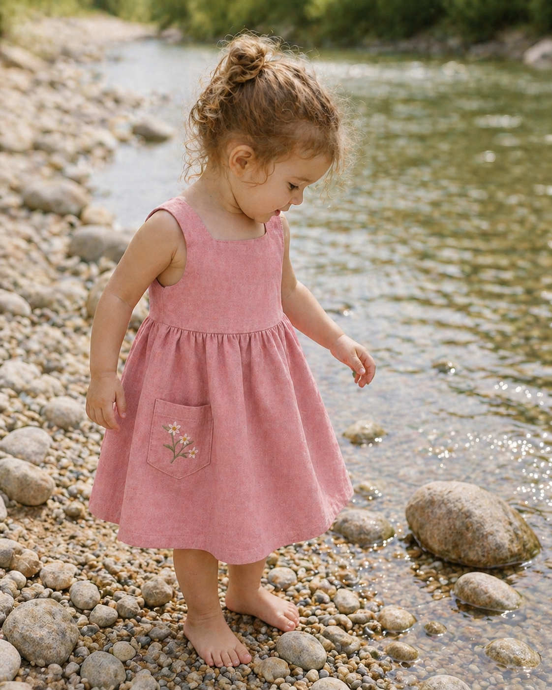 Young girl in a pink dress standing by a stream on a pebbly shore.